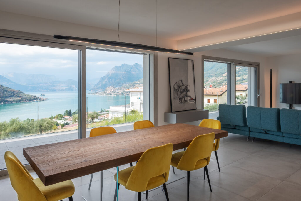 fotografia interno soggiorno moderno open space con divano blu tavolo legno sedie gialle illuminazione LED integrata nelle pareti vista lago iseo laura farina architetto